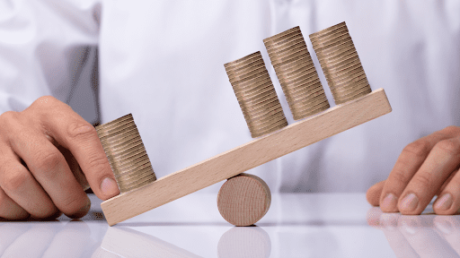 Businessperson's Hand Showing Unbalance Between Stacked Coins On Wooden Seesaw