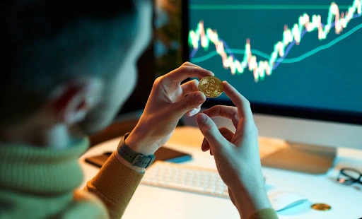 a man looking at a bitcoin on his hand, with a trading chart on a computer monitor as background