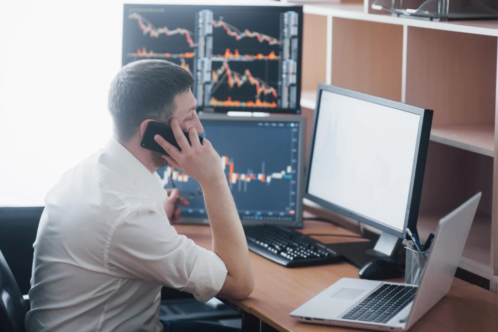 man looking at monitors with trading charts while making a call with a phone